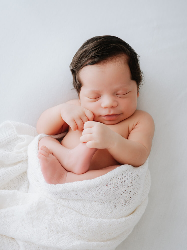 Baby boy wrapped in blanket holding foot - photographed by Natasha Cranley